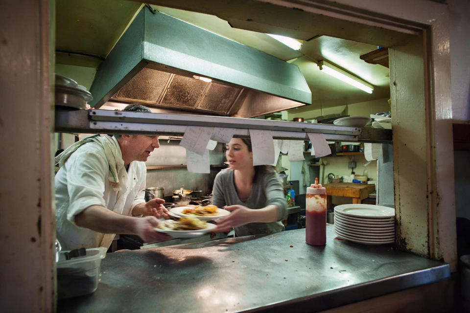 A cook passing two plates to a server through the kitchen hatch of a small restaurant